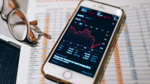 Close-up of smartphone with stock market graph on table with financial documents and glasses.