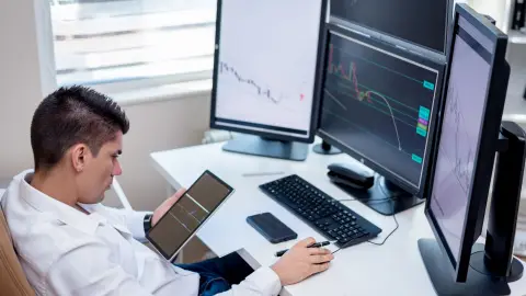 Business professional analyzing financial charts on monitors and tablet in modern office workspace.